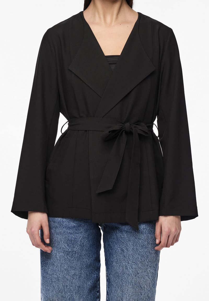 Veste enveloppante noire à manches larges, taille ceinturée et texture lisse. Présente un col en V profond et des poches à l'avant. Associée à un jean bleu.