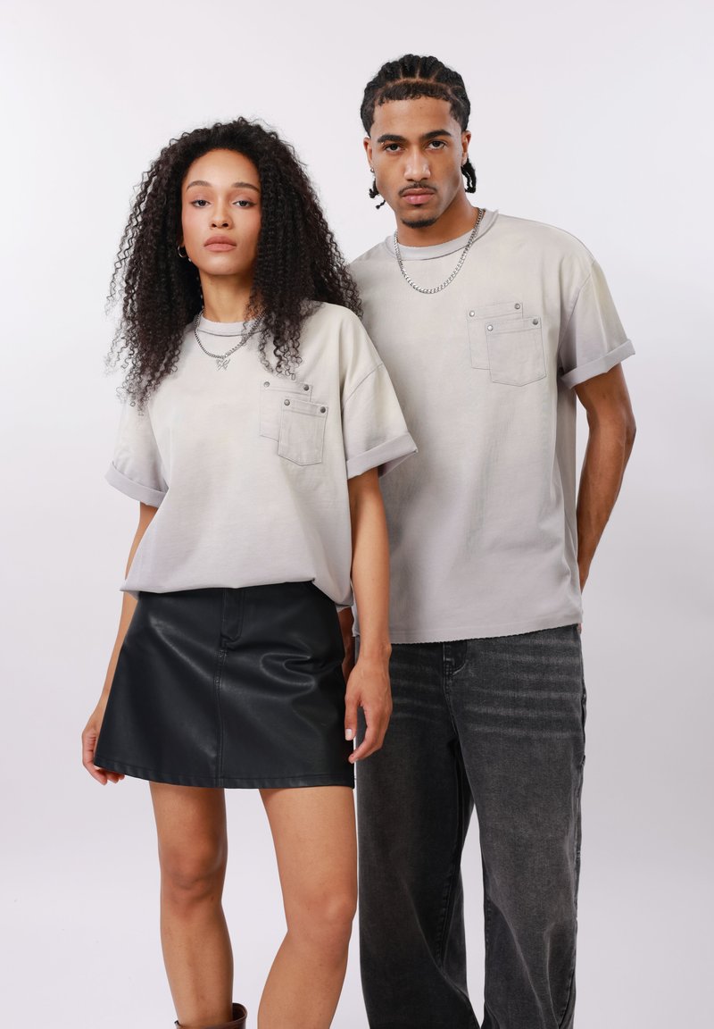 Γυναίκα με σγουρά μαλλιά φοράει ανοιχτό γκρι μπλουζάκι με τσέπες και μαύρη δερμάτινη φούστα. Άνδρας με πλεκτά μαλλιά φοράει αντίστοιχο μπλουζάκι και σκούρο τζιν.