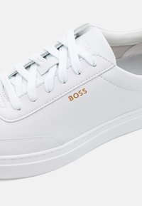 White sneaker with a smooth leather finish, featuring light blue laces, embossed 'BOSS' logo in gold, and a simple, streamlined design.