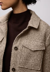 Béžová texturovaná bunda s chlupatým povrchem, vybavená dvěma náprsními kapsami a velkými tmavými knoflíky. N nošena přes tmavě hnědý žebrovaný rolák.