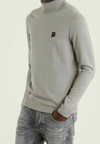 Pull à col roulé gris clair en tissu doux, avec un patch logo noir sur la poitrine et des poignets et un ourlet côtelés.