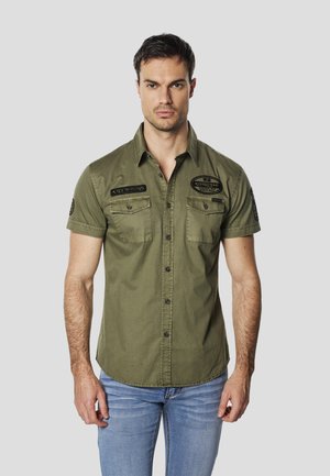 Rövid ujjú olívazöld pamuting, fekete foltokkal, két mellzsebbel és gombos elülső résszel.