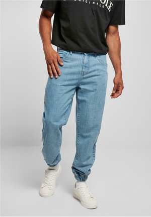 Mann trägt hellblaue Jogging-Jeans aus Denim mit elastischen Bündchen, weiße Sneaker, weiße Socken und ein schwarzes T-Shirt mit weißem Text.
