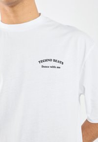 Balta medvilninė marškinėliai su juodu spausdintu tekstu „TECHNO BEATS“ ir „Šok su manimi“ kreivame išdėstyme. Trumpi rankovės ir apvalus kaklo dizainas.