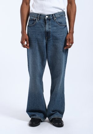 Faded blue denim jeans, wide leg design, five-pocket style, logo accents on the back, with a slight fraying at the hem.