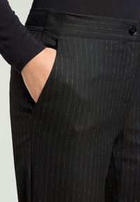 Zwarte broek met krijtstreep en een gladde textuur, met gouden strepen, zijzakken en een zwarte knoop aan de tailleband.