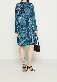 Long-sleeve dress in navy with teal and turquoise paisley print, flared hem, paired with black heeled sandals and a small black handbag.