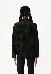 Camisa de manga longa, com collar e punhos com botão, de cor verde escuro e com riscas, combinada com calças pretas, mostrando a vista traseira. Textura lisa.