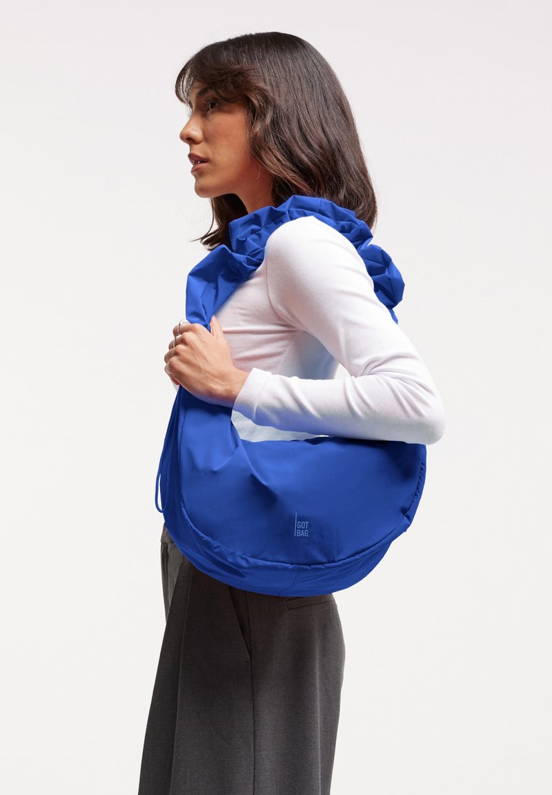 Borsa a spalla in tessuto blu con parte superiore arricciata, texture morbida e dettaglio del logo. Indossata da una persona con una maglietta aderente bianca e pantaloni grigi.