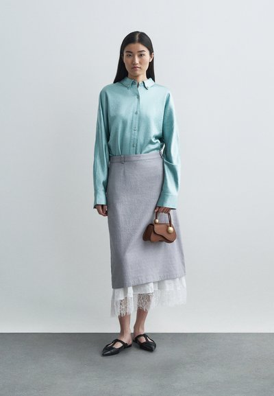 Nainen pukeutuneena vaaleansiniseen nappipaitaan, harmaaseen kietaisuhameeseen valkoisen pitsihameen yli, mustiin teräväkärkisiin ballerinoihin, pitäen pientä ruskeaa käsilaukkua.