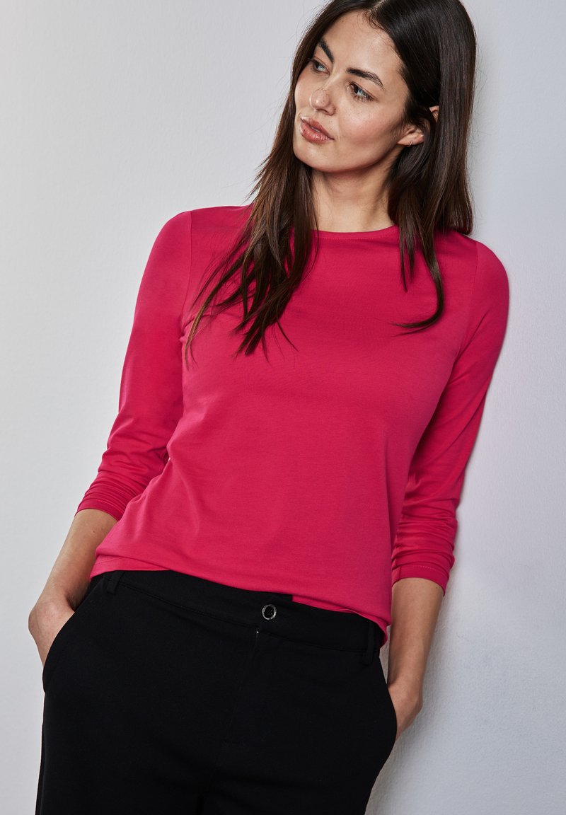 Maglietta attillata a maniche lunghe in tessuto fucsia brillante, con scollatura rotonda, abbinata a pantaloni neri con tasca frontale e dettaglio in bottone.