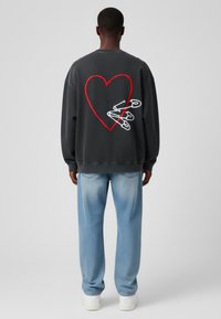 Sudadera gris con un gran corazón rojo y alfileres de seguridad blancos bordados en la parte posterior, combinada con jeans de un azul claro y zapatillas blancas.