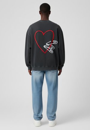 Felpa grigia con un grande cuore rosso e spille di sicurezza bianche ricamate sulla schiena, abbinata a jeans azzurri chiari e sneaker bianche.