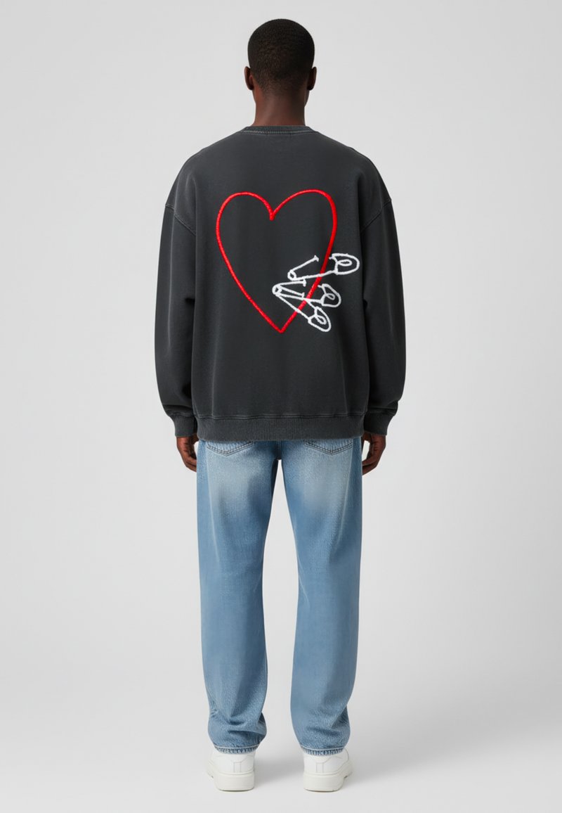 Sudadera gris con un gran corazón rojo y alfileres de seguridad blancos bordados en la parte posterior, combinada con jeans de un azul claro y zapatillas blancas.