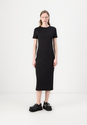 Schwarzes T-Shirt-Kleid in knie-langer Länge mit kurzen Ärmeln, aus weichem Material gefertigt. Kombiniert mit klobigen schwarzen Plateauschuhen. Minimalistisches Design.