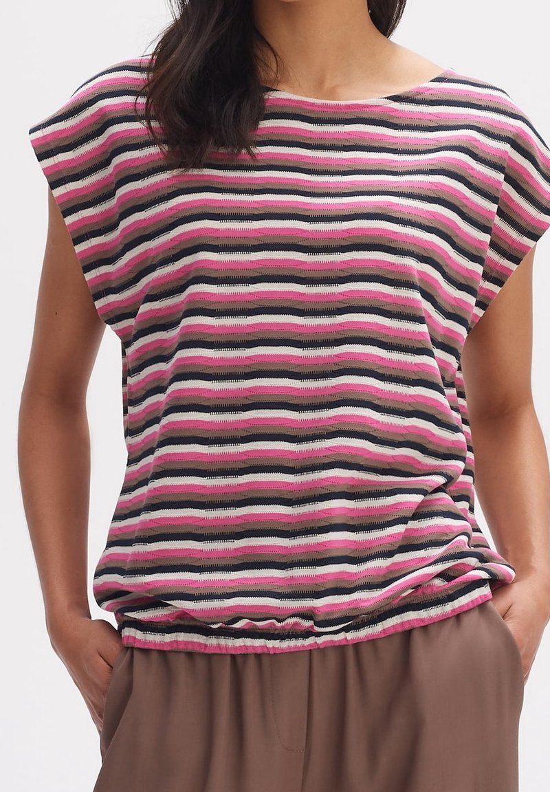 Femme portant un top court en maille à manches courtes avec des rayures horizontales roses, noires, blanches et grises, rentré dans un pantalon marron.