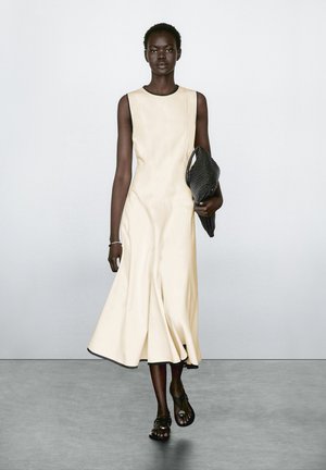 Cremfarbendes ärmelloses Kleid mit weit auslaufendem Saum, das eine glatte Textur und einen dunklen Besatz aufweist. Das Model hält eine strukturierte schwarze Clutch und trägt dunkle Sandalen.
