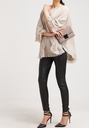 Vrouw in een beige franje poncho, zwarte nepleer legging, zwarte puntige hoge hakken, met een gestoffeerde taupe clutchtas.