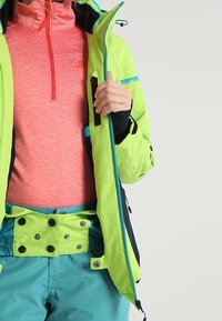 Světlá zelená nepromokavá bunda s modrými akcenty, vrstvená přes růžovou mikinu na zip. Bunda má zipovou kapsu a kapsy na patentky.