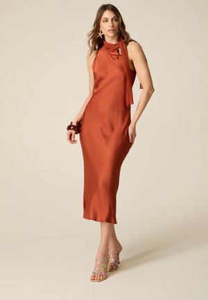 Donna con un abito midi in satin senza maniche color ruggine con un fiocco al collo, grandi orecchini, braccialetti e sandali con tacco alto e cinturini.
