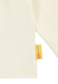 Cream cotton t-shirt featuring long sleeves, a rounded neckline, and a rectangular yellow tag with red writing near the bottom left.