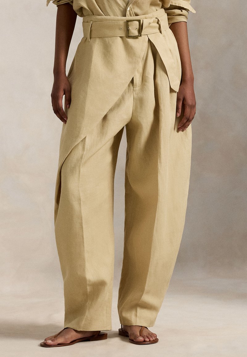 Μπεζ λινό παντελόνι με ψηλή μέση και φαρδιά παντελόνα, με ζώνη στη μέση και ασύμμετρα πιέτες για επιπλέον υφή και λεπτομέρεια.