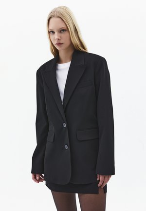 Ung kvinde med langt blondt hår iført en oversized sort blazer, hvid skjorte, sort mininederdel og gennemsigtige sorte strømpebukser foran en hvid baggrund.