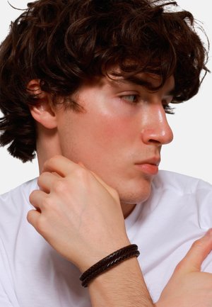 Brown leather braided bracelet worn on the wrist, features a smooth finish, paired with a plain white T-shirt. Profile view of a male model.