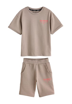 Chico adolescente de pie con una mano en el bolsillo, vistiendo camiseta beige oversize y pantalones cortos, calcetines blancos de marca y zapatillas deportivas sobre fondo gris.