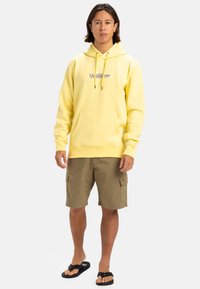 Sudadera amarilla con un bolsillo frontal, cordones ajustables y logo. Combinada con pantalones cortos de cargo color caqui y chanclas negras.