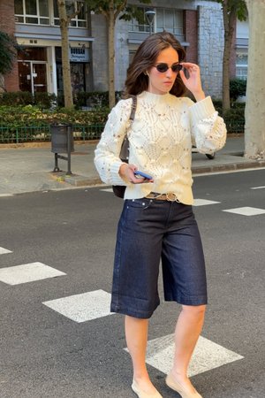 Crème gebreide trui met textuurpatronen, gecombineerd met donkere denim culottes. Beigekleurige flats en een zonnebril maken de look compleet.