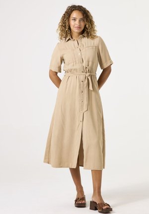 Vestido camel con botones y mangas cortas, cinturón en la cintura y dos bolsillos en el pecho. Tejido suave con largo midi y una abertura central.