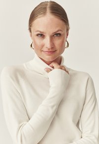 Weißer Rollkragenpullover mit langen Ärmeln, geripptem Kragen und glatter Textur. Das Model hält zart die Finger in der Nähe des Ausschnitts vor einem neutralen Hintergrund.
