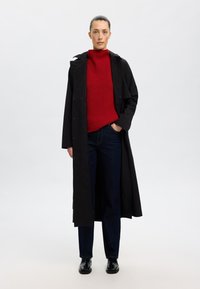 Schwarzer Trenchcoat über einem roten gerippten Pullover, kombiniert mit dunklen Jeans und schwarzen Knöchelstiefeln. Der Mantel hat einen Kragen und Knöpfe.