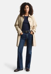 Beige trench coat with large buttons over a navy blue top, paired with dark blue flared jeans and brown leather boots.