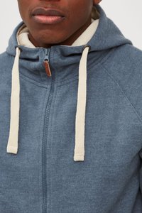 Blå zip-up hettegenser med en myk tekstur, med beige snorer og en liten brun merkelapp ved glidelåsen.