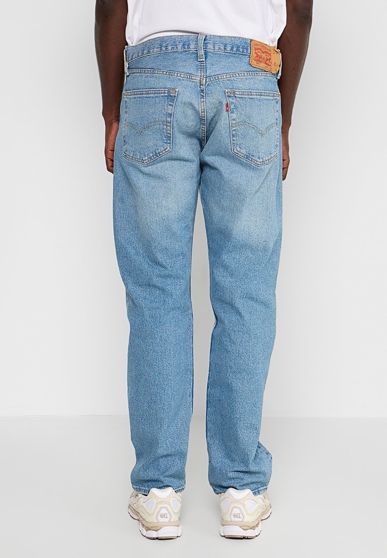 Mann in hellblauen geraden Jeans und weißen Sneakers, mit entspannten Händen an den Seiten, von hinten vor weißem Hintergrund gezeigt.