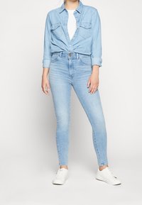 Jeans ajustados de denim azul claro con cintura alta y un suave desgaste. Combinados con una camisa de denim abotonada y zapatillas blancas.