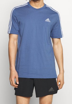Mężczyzna w niebieskiej koszulce Adidas z białymi paskami na rękawach oraz czarnych spodenkach Adidas, stojący na jednolitym tle.
