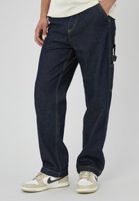 Pantalons en denim bleu foncé avec des surpiqûres jaunes, design à jambe droite et détails de poches latérales. Associés à des baskets beige et blanches.