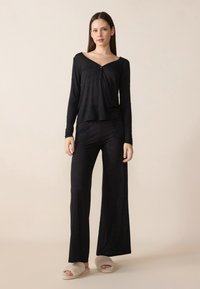 Μαύρη μπλούζα με μακριά μανίκια και V-λαιμό, μ ε λεπτομέρεια κουμπιού και μαύρο παντελόνι σε ελεύθερη γραμμή, φτιαγμένο από μαλακό ύφασμα; σε συνδυασμό με ανοιχτόχρωμες, αφράτες παντόφλες.