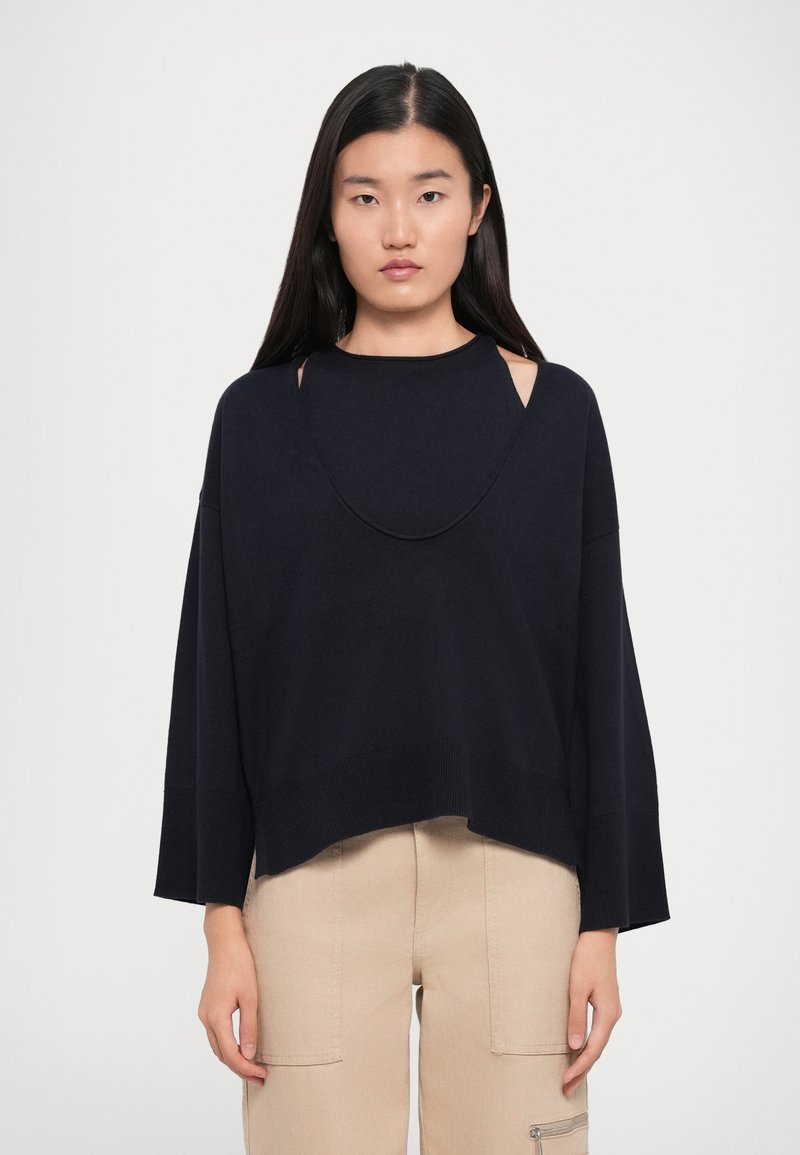 Maglione cropped blu navy con scollatura ampia e dettagli ritagliati. Materiale morbido, vestibilità ampia e bordo a costine.