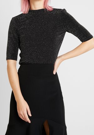 Persona con pelo rosa que lleva un top negro brillante de manga corta y una falda negra de talle alto con una abertura frontal, mano en la cadera.