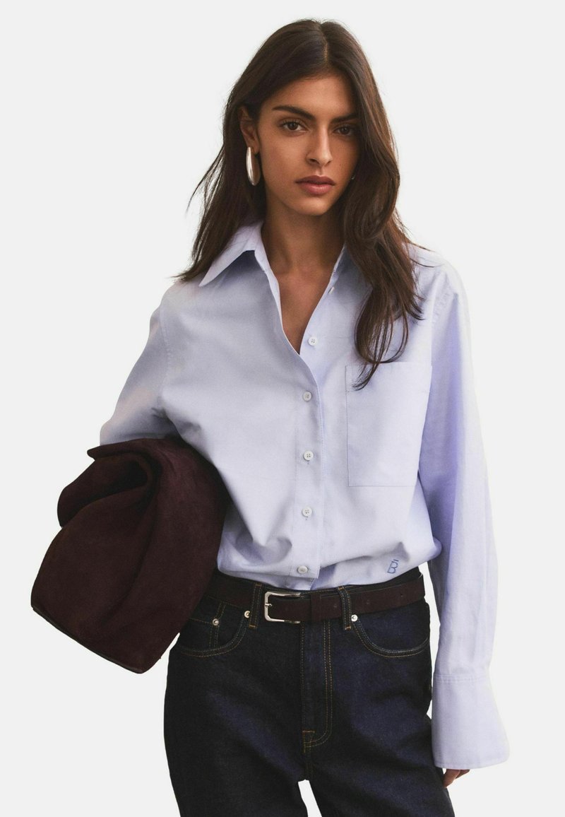 Mujer con cabello largo y oscuro, vistiendo una camisa azul claro abotonada y jeans oscuros, sosteniendo una chaqueta marrón oscuro doblada sobre su brazo.