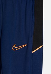 Pantalons de sport marine avec des accents noirs, présentant un logo Nike brodé orange. Fabriqués à partir d'un matériau lisse et léger.