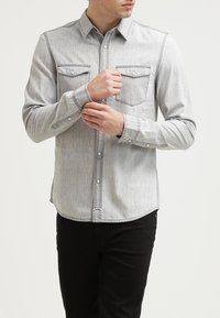 Chemise boutonnée gris clair avec deux poches frontales, coutures contrastées et manches longues, associée à un pantalon noir.