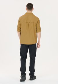 Bruine blouse met opgerolde mouwen en een rugplooi, gedragen met zwarte lichte broek en zwarte schoenen, zicht vanuit de rug.