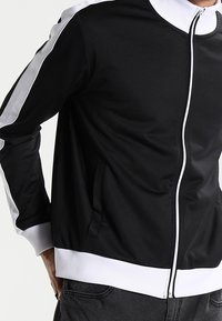 Black zip-up jacket with white accents and ribbed cuffs. Features a high collar and side pockets. Made from smooth, flexible fabric.