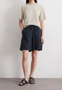 Persona che indossa una maglietta beige a maniche corte, pantaloni corti blu scuro con cordino larghi e sandali neri con fibbie dorate, in piedi contro un muro bianco.