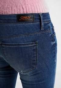 Jeans azul oscuro con un diseño ajustado, que presentan bolsillos traseros y una etiqueta en la cintura. El material parece ligeramente desgastado y tiene textura.